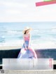 A woman in a pink dress and straw hat sitting on a wall by the ocean.