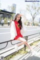 A woman in a red dress and white jacket sitting on a bench.